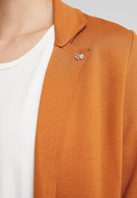Blazer arancione in tessuto texturizzato, con un'accattivante accentuazione a fiocco argento sul bavero. Indossato su una maglietta bianca.