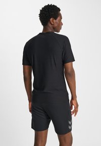 Man in een zwart shirt met korte mouwen en zwarte korte broek met reflecterende chevronstrepen, staand met de rug naar voren tegen een effen witte achtergrond.