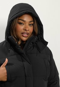 Czarna kurtka puchowa z kapturem, z zamkiem błyskawicznym i przednią kieszenią. Materiał ma gładką teksturę z pikowanym przeszyciem.