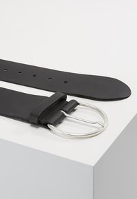 Ceinture en cuir noir dotée d'une boucle en métal argenté arrondie, de plusieurs trous d'ajustement et d'une surface légèrement texturée.