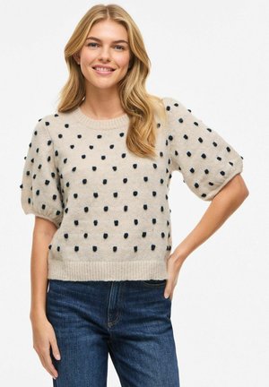 Smiling woman with long blonde hair wearing a beige short-sleeve sweater with black polka dots and dark blue jeans, hand on hip.