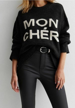 Zwarte trui met witte tekst "MON CHÉR", gedragen met zwarte hooggetailleerde broek en een zwarte riem met zilveren gesp, handen van het model ontspannen langs het lichaam.