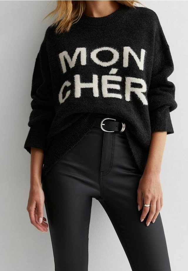 Pull noir avec texte blanc "MON CHÉR", porté avec un pantalon noir taille haute et une ceinture noire à boucle argentée, mains du modèle relâchées le long du corps.