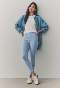 Blugi skinny bleu deschis, tricou alb și o cămașă supradimensionată cu dungi albastre, asortate cu pantofi crem cu șireturi pe un fundal neutru.