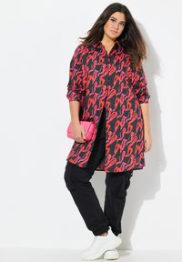 Abito camicia nero con un vivace motivo astratto rosso e rosa; presenta una chiusura frontale con bottoni, maniche lunghe e una vestibilità ampia, abbinato a pantaloni neri.