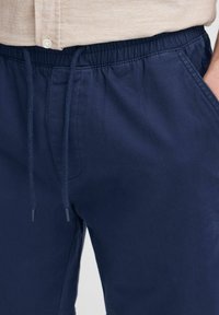 Pantalone tamnoplave boje s vezicom u struku i bočnim džepovima; glatka tkanina s suptilnim sjajem i pojačanim šavovima.