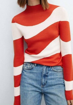 Personne portant un pull à manches longues et col montant avec de larges rayures diagonales orange vif et blanches, associé à un jean bleu en denim.