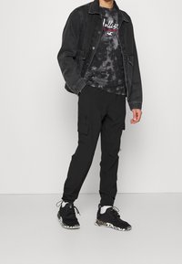 Svart denimjacka över en svart och grå tie-dye t-shirt, kombinerad med svarta cargobyxor och svarta sneakers med en kamouflagemönstrad sula.