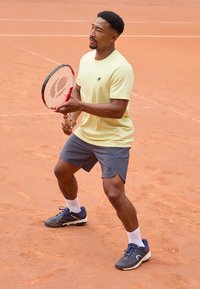 Gul atletisk skjorte med logo, grå shorts og marineblå tennissko. Holder en rød og hvit tennisracket på leircourt.