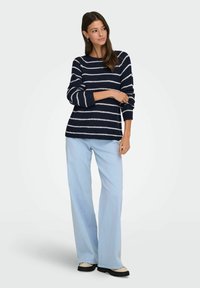 Pull en laine rayé bleu marine et blanc avec un col rond et des manches longues, assorti à un pantalon bleu clair à jambes larges et des chaussures à semelles noires.