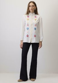 Bela dolga majica z dolgimi rokavi z barvitim cvetličnim vezenjem spredaj, nagubanim ovratnikom in gumbi. Ujemite jo z črnimi širokimi hlačami.