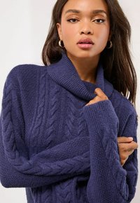 Granatowy sweter z splotem w warkocze z podwiniętym kołnierzem, teksturowanym materiałem i długimi rękawami, charakteryzujący się luźnym krojem i widocznymi detalami szwów.