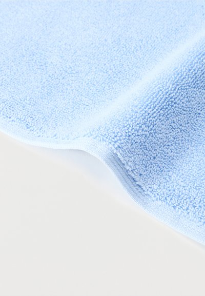 Πετσέτα από περκάλι σε ανοιχτό γαλάζιο με ορατή υφή βρόχων, μερικώς διπλωμένη πάνω σε λευκή επιφάνεια.