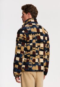 Giacca in pile multicolore con un motivo geometrico, caratterizzata da tonalità di blu, marrone, crema e nero, con un design a colletto alto.