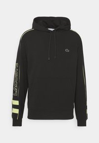 Svart hoodie med snörning, framficka och Lacoste-logotyp. Har neongula ränder längs ärmarna för kontrast. Tillverkad av bomullsmix.