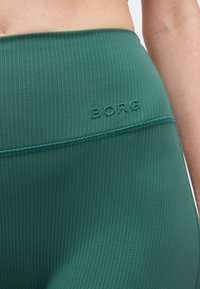 Κοντινό πλάνο πράσινων ριμπ λεπτομερειών κολάν με ψηλή μέση και το λογότυπο "BORG" που είναι ανάγλυφο κοντά στη μέση στο ισχίο ενός ατόμου.