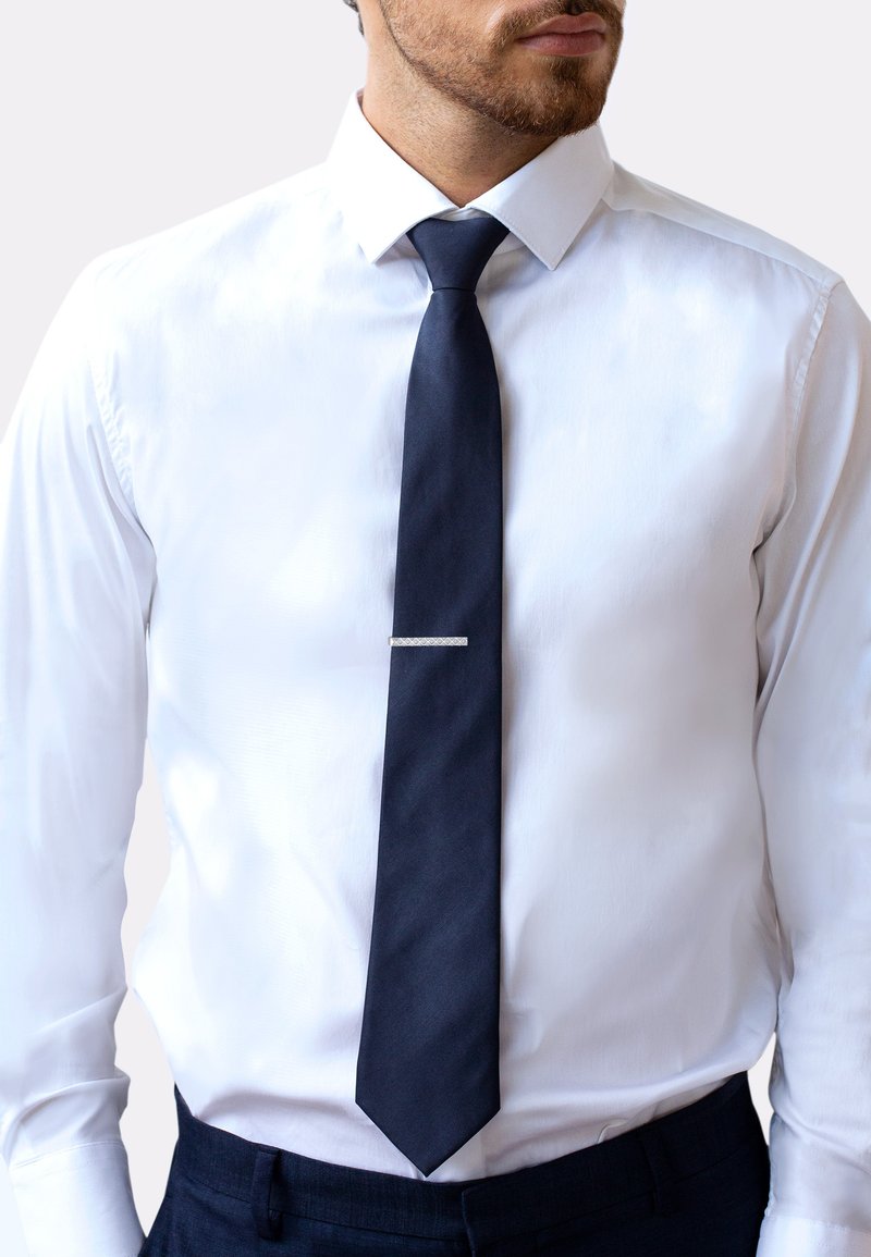 Cravate bleu marine avec une texture lisse et une pointe, ornée d'un détail en clip métallique sur le devant, associée à une chemise habillée blanche.