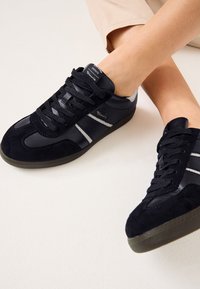Baskets noires avec texture en daim et cuir, dotées de lacets plats, de rayures latérales grises et d'une semelle en caoutchouc. Logo de la marque sur la languette.