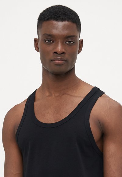 Schwarzes Baumwoll-Tanktop mit einem runden Halsausschnitt und weiten Armausschnitten, das eine taillierte Passform und eine glatte Textur aufweist. Das Modell hat einen kurz geschnittenen Haarschnitt.