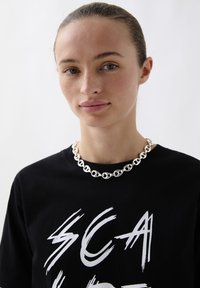 Zwarte T-shirt met witte tekst, gecombineerd met een zilveren kettinghalsketting bestaande uit onderling verbonden cirkelvormige stukken, gladde oppervlakte en ronde randen.