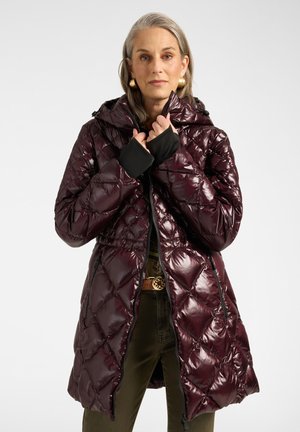 Mujer con un abrigo acolchado de color púrpura oscuro brillante con capucha, mangas interiores negras, pantalones verdes y pendientes dorados sobre un fondo liso y claro.