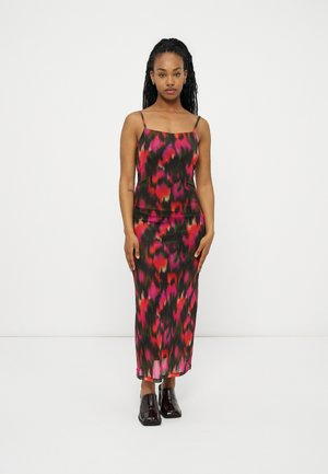 Donna con lunghi capelli neri che indossa un abito lungo colorato senza maniche e stivaletti scuri alla caviglia, in piedi contro uno sfondo bianco.