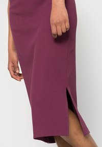 De hand van een vrouw rustend bij de zijsplit van een geribbelde, knielange, pruimenpaarse rok.