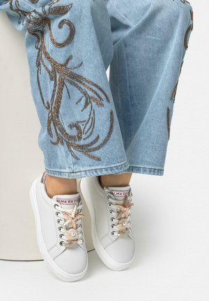 Zapatillas blancas con adornos de conchas marinas, estrella de mar y perlas en los cordones, usadas con jeans azul claro que presentan bordados marrones decorativos.