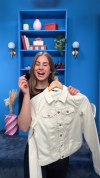 Kvinde holder beige denimjakke på bøjle i blåt rum med hylder, planter og dekorative genstande, smilende og viser stofprøve.