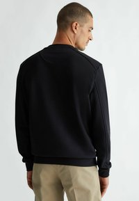 Felpa nera con collo a giro, polsini a costine e una texture liscia, abbinata a pantaloni beige, vista da dietro.