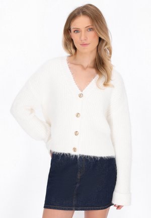 Witte, fluffy gebreide cardigan met een V-hals, vijf decoratieve gouden knopen en geribbeld patroon. Gecombineerd met een donkere denim mini-rok.