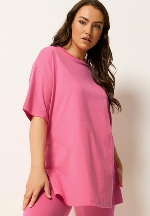 Kvinde med langt brunt hår iført løs lyserød kortærmet skjorte og matchende lyserøde bukser, stående foran en ensfarvet lys baggrund.