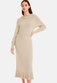 Beiges Strickkleid mit gerippter Textur, das einen Rüschenausschnitt und einen ausgestellten Saum aufweist. Lange Ärmel und figurbetonte Silhouette.