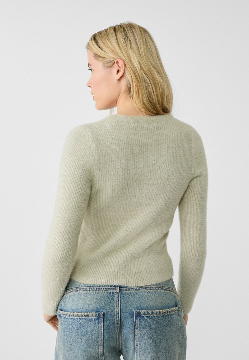 Hellgrüner, gestrickter Pullover mit weicher Textur, schmaler Passform und rundem Ausschnitt, kombiniert mit hochgeschnittenen blauen Jeans mit Gesäßtaschen.