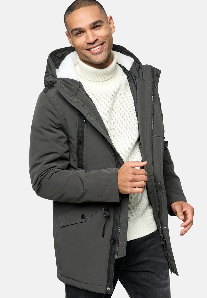 Grå isoleret parka med fleece-foret hætte, knap- og lynlåslukninger samt sidelommer, båret over en struktureret hvid sweater.