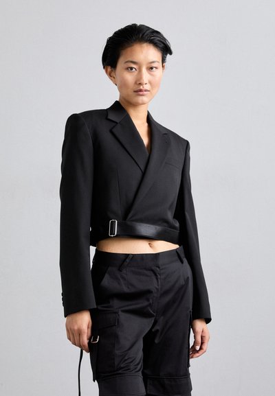 Blazer noir court avec un décolleté en V profond, des épaules structurées et une ceinture à la taille. Associé à un pantalon cargo noir avec des poches latérales.