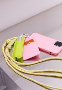 Funda de teléfono de silicona rosa, portatarjetas verde y objeto de metal, todos con un cordón trenzado que presenta detalles en patrón rosa y verde.