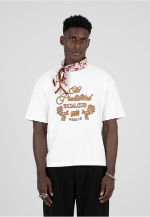 White t-shirt featuring "The Prohibited Social Club" in gold lettering, flanked by lion graphics; paired with a floral-patterned scarf.