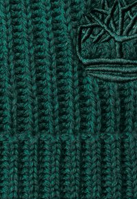 Grüne gestrickte Mütze mit vertikaler Rippstruktur. Verfügt über ein gesticktes Baum-Logo in einem dunkleren Grünton. Warme und gemütliche Gestaltung.