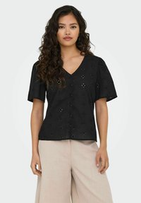 Schwarze Baumwollbluse mit V-Ausschnitt und kurzen Ärmeln. Mit Spitzenverzierungen und strukturierten Knöpfen, in lockerer Passform, kombiniert mit hellbeigen Hosen.