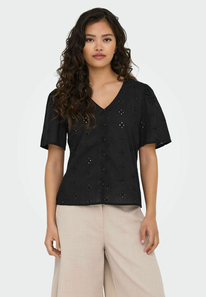 Schwarze Baumwollbluse mit V-Ausschnitt und kurzen Ärmeln. Mit Spitzenverzierungen und strukturierten Knöpfen, in lockerer Passform, kombiniert mit hellbeigen Hosen.