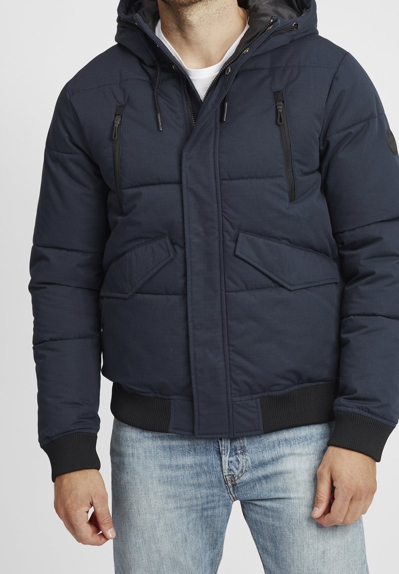 Granatowa ocieplana kurtka z kapturem, zamkiem błyskawicznym z przodu, dwoma bocznymi kieszeniami oraz dwoma kieszeniami na piersi zapinanymi na zamek. Ściągacze na mankietach i dole kurtki.