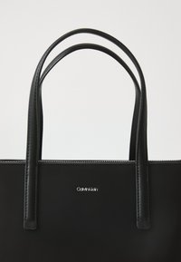 Borsa tote in pelle nera con due manici allungati. Presenta una cucitura bianca lungo il bordo superiore e un logo Calvin Klein argentato sul davanti.