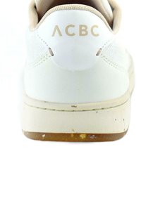 Zapatilla blanca con un upper texturizado, acento crema alrededor del talón y el logo "ACBC" en dorado. Ligero desgaste en la suela de goma color tostado.