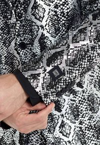 Schwarze und silberne gemusterte Blazer mit Schlangenhautdesign, geknöpften Manschetten und einem Logoaufnäher am Ärmel. Sichtbare Details aus strukturierter Stoff.