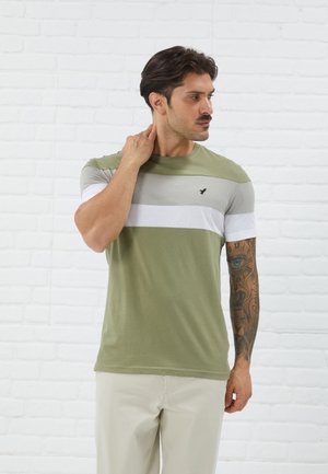 T-shirt rayé vert olive et gris à manches courtes, avec un logo noir sur la poitrine, assorti à un pantalon de couleur crème.