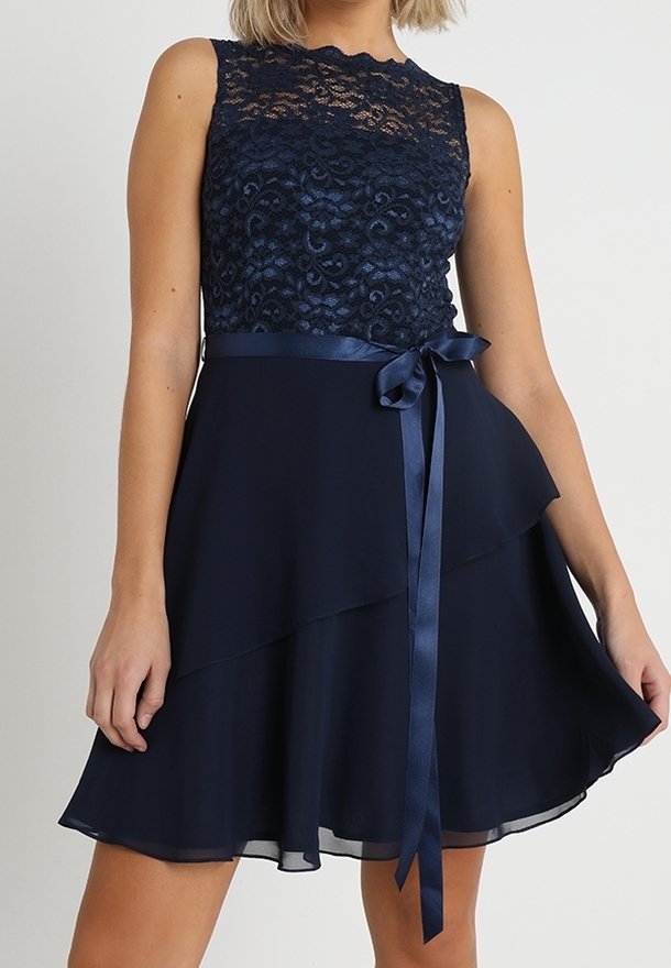 Robe sans manches bleu marine avec corsage en dentelle, ruban en satin à la taille noué en nœud et jupe en chiffon à plusieurs couches portée par une personne debout.