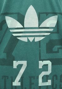 Logo trefoil Adidas blanc au-dessus de grands chiffres blancs "72" sur un fond en tissu vert avec un texte graphique sombre et estompé.
