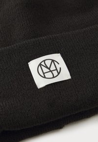 Bonnet noir en tricot avec une étiquette carrée blanche. L'étiquette affiche un logo circulaire avec des lettres stylisées. La texture semble douce et extensible.