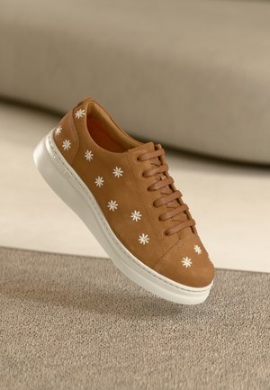 Brauner Sneaker aus Wildleder mit weißer Blumenstickerei, rundem Zehenbereich, Schnürsenkeln und weißer Gummisohle. Schwebt über einer strukturierten Oberfläche.
